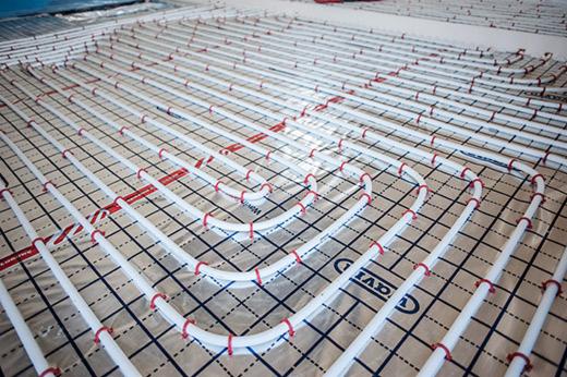 Białe rury ogrzewania podłogowego ułożone na folii z nadrukowaną siatką, spięte czerwonymi uchwytami. Widok z góry, instalacja przed wylewką.