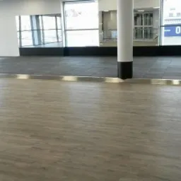 Wykończona podłoga z paneli imitujących drewno przechodząca w szarą wykładzinę kwadratową, oddzielona metalową listwą w przestrzeni biurowej.