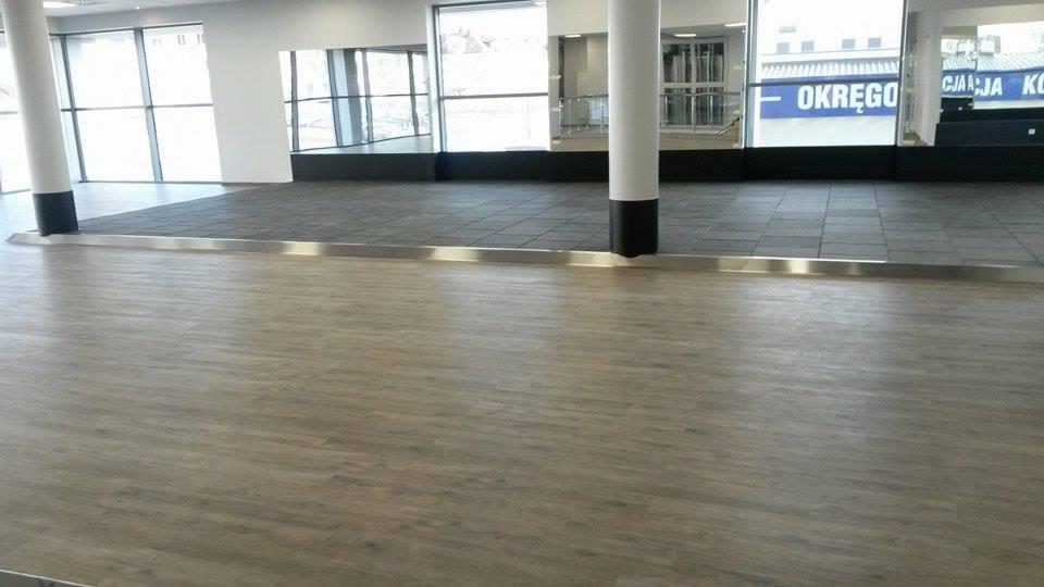 Wykończona podłoga z paneli imitujących drewno przechodząca w szarą wykładzinę kwadratową, oddzielona metalową listwą w przestrzeni biurowej.