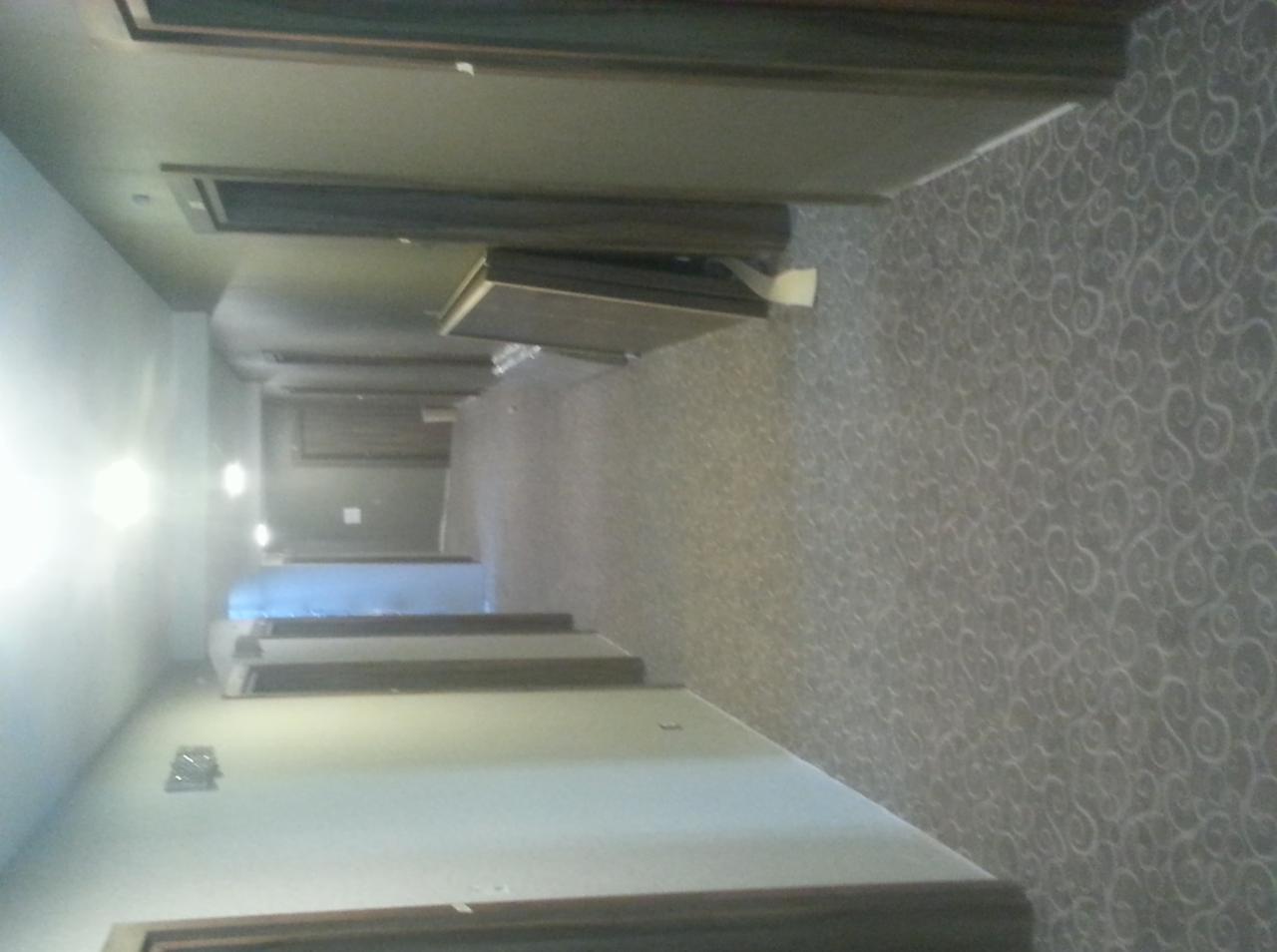 Długi korytarz z wzorzystą wykładziną podłogową w odcieniach szarości, widoczne drzwi i oświetlenie. Ułożona wykładzina w obiekcie komercyjnym.