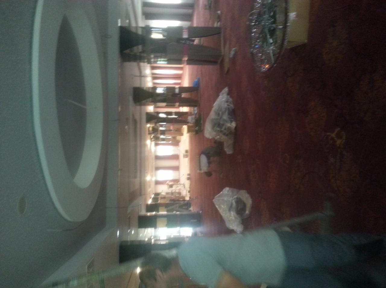 Montaż czerwonej wykładziny w przestronnym wnętrzu z dekoracyjnym sufitem. Rozpakowane elementy oświetlenia czekają na instalację.