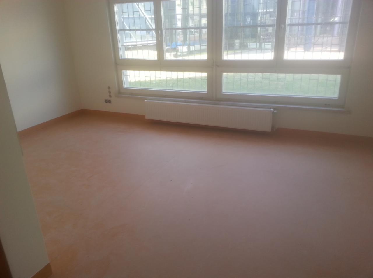 Pokój po położeniu jednolitej, pomarańczowej podłogi z tworzywa sztucznego. Widoczne trzy okna z kratami i biały grzejnik pod nimi. Świeże wykończenie wnętrza.