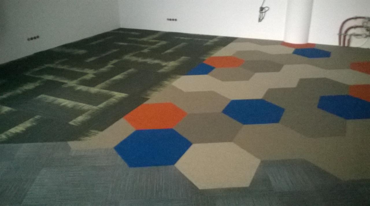 Ułożona wykładzina dywanowa w geometryczne wzory: heksagony w kolorach beżowym, niebieskim i pomarańczowym oraz wzór w ciemnych odcieniach zieleni.