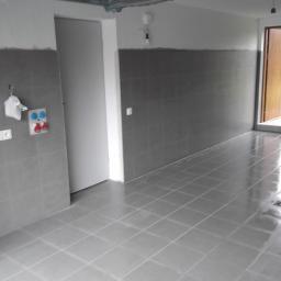 Art-bud Artur Wrzoszczyk - Wykończone płytki podłogowe i ścienne w szarym odcieniu, z widoczną instalacją elektryczną i sanitarną na ścianie. Drzwi wejściowe w tle, po prawej stronie.