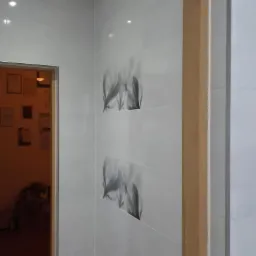 Białe płytki ścienne z motywem liści w kształcie rombu, drewniana rama drzwiowa, widok na pomieszczenie z obrazami na ścianie. Wykończenie wnętrza.