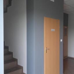 Art-bud Artur Wrzoszczyk - Korytarz po malowaniu: szare ściany, drewniane drzwi z metalową klamką, schody. Widoczne detale wykończenia i odświeżone kolory.