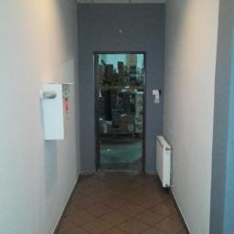 Art-bud Artur Wrzoszczyk - Wnętrze korytarza z brązową podłogą w romby, białą gaśnicą na ścianie i grzejnikiem. W tle widoczne otwarte drzwi do magazynu.