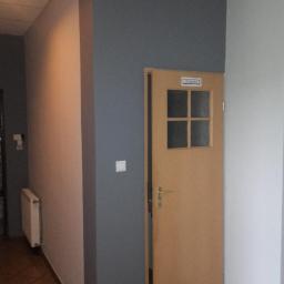 Art-bud Artur Wrzoszczyk - Ściana pomalowana na dwa kolory, szary i jasny beż, z drewnianymi drzwiami do toalety po prawej stronie. Widoczny fragment korytarza z grzejnikiem.