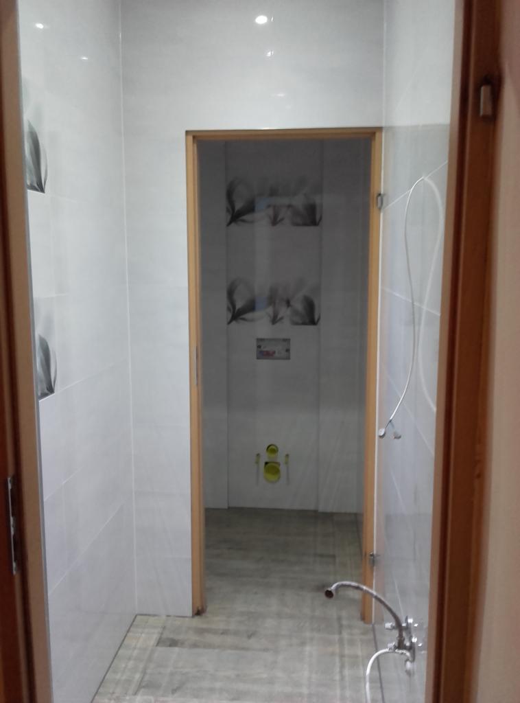 Wykończona łazienka z białymi płytkami, dekoracyjnymi akcentami i zamontowaną instalacją sanitarną. Widok z zewnątrz, z otwartymi drewnianymi drzwiami.