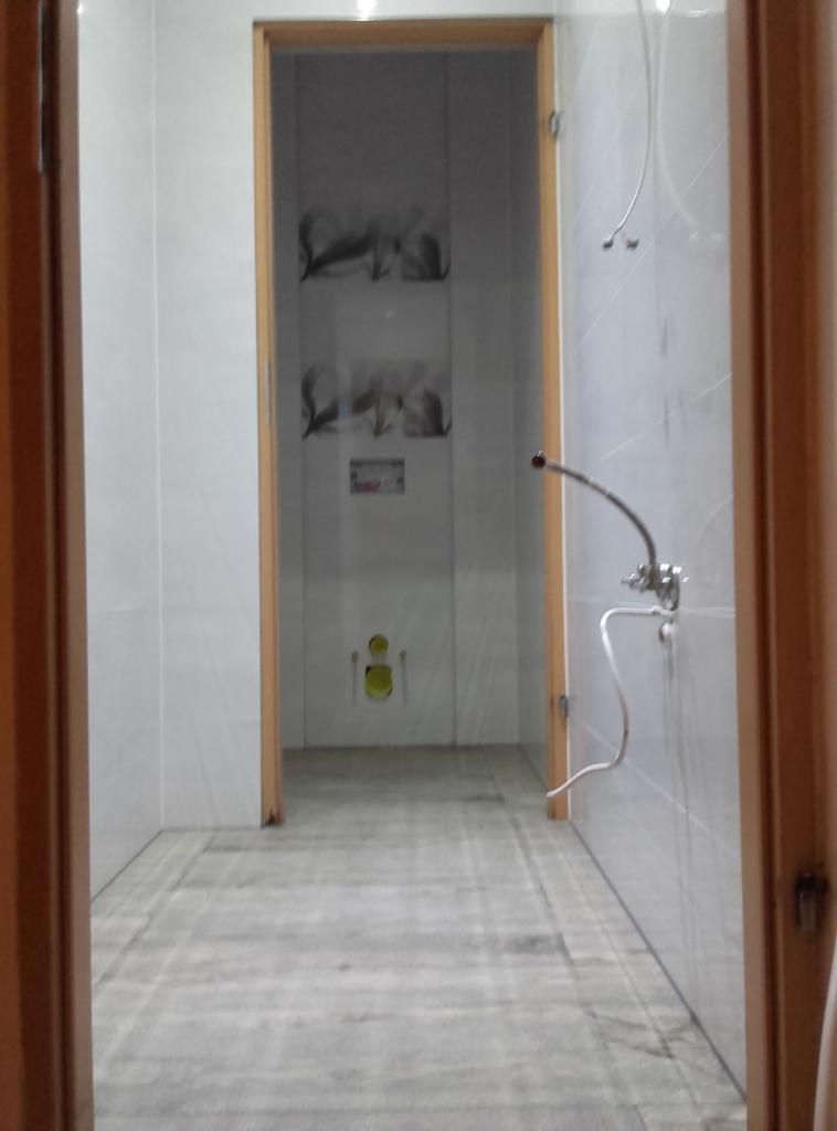 Wykończona łazienka z szarymi płytkami podłogowymi i białymi ściennymi. Widoczne przyłącza do armatury i fragment dekoru na ścianie w głębi pomieszczenia.