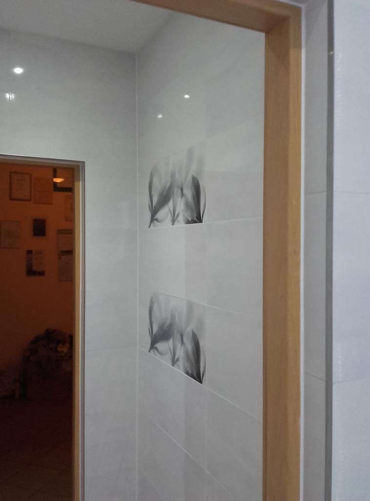 Białe płytki ścienne z motywem liści w kształcie rombu, drewniana rama drzwiowa, widok na pomieszczenie z obrazami na ścianie. Wykończenie wnętrza.