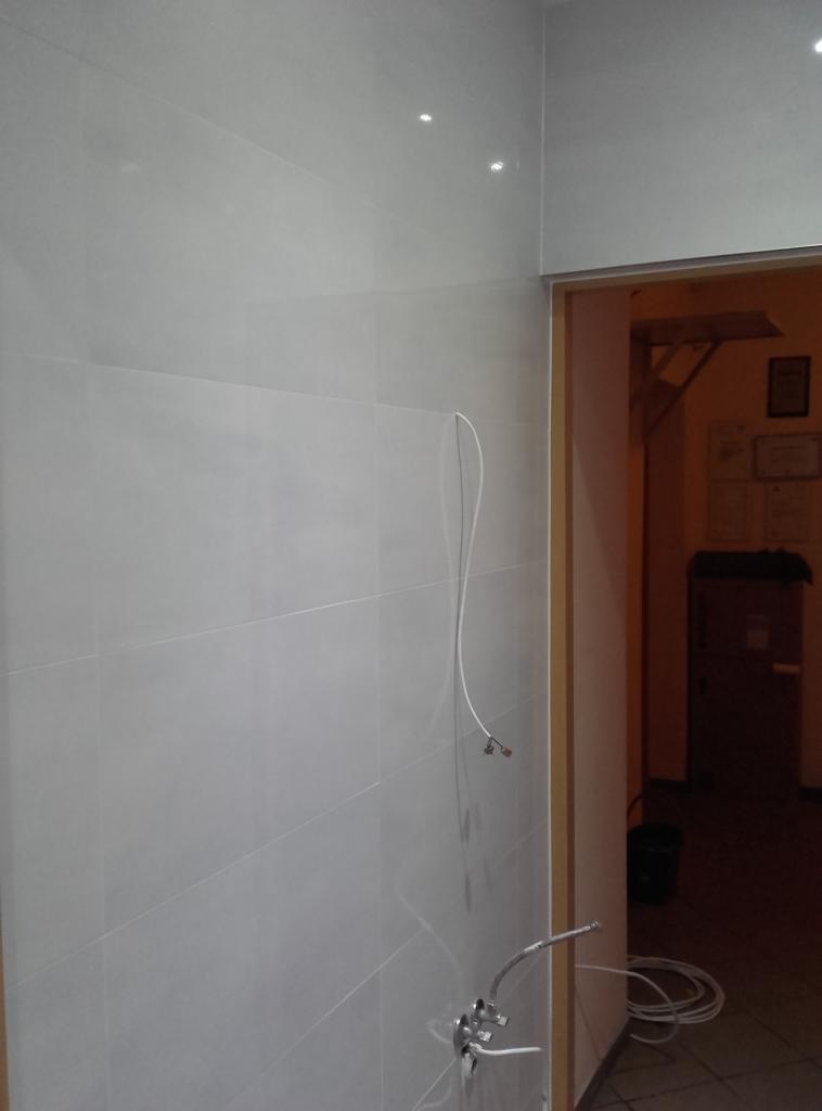 Ściana wyłożona szarymi, podłużnymi płytkami w łazience. Widoczne wystające kable i rurki instalacji wodnej przed montażem armatury. W tle widoczne otwarte drzwi.