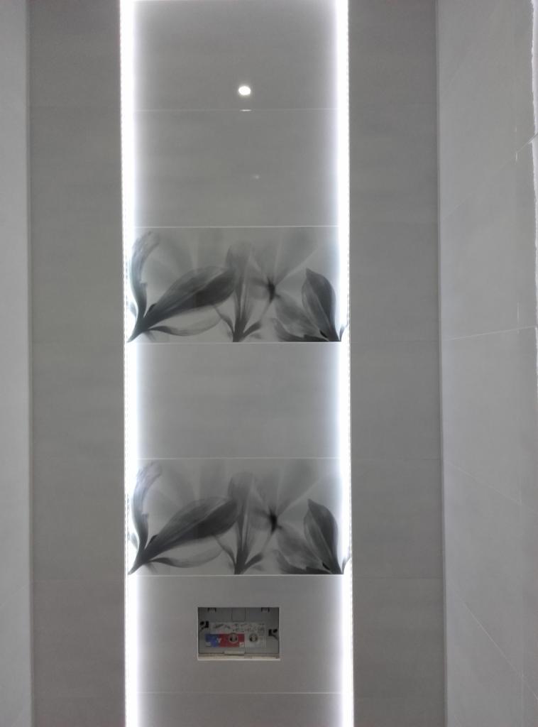 Wąski pas płytek z motywem kwiatowym, podświetlony pionową listwą LED, wkomponowany w jasną ścianę. Widoczna skrzynka z instalacją elektryczną.