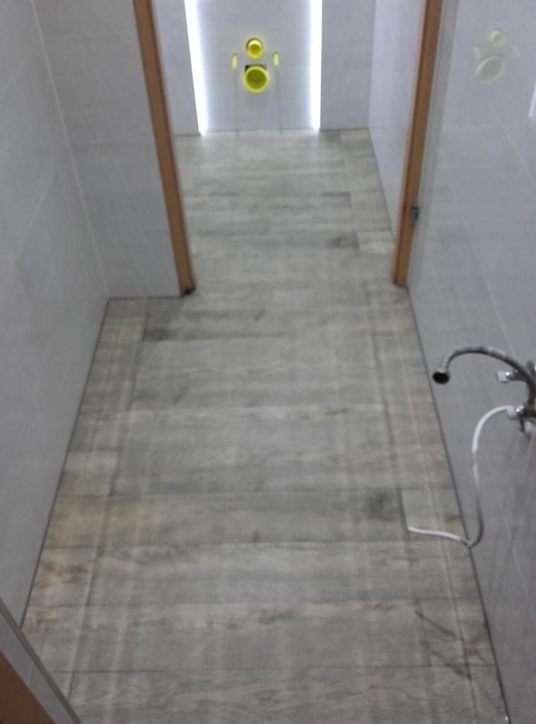 Wykończona łazienka z szarą podłogą imitującą drewno i białymi ścianami. Widoczne elementy armatury i oświetlenia. Ukończony remont łazienki.