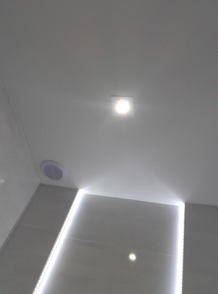 Biały sufit z kwadratową lampą LED i okrągłym nawiewnikiem wentylacyjnym. Ściany z podświetleniem LED tworzą nowoczesny design wnętrza.