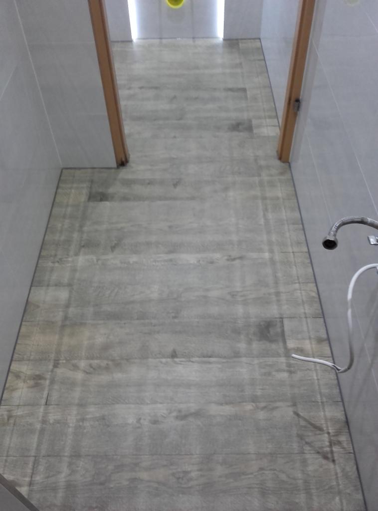 Ułożone panele podłogowe imitujące drewno w wąskim korytarzu z szarymi ścianami i widoczną instalacją wodną po prawej stronie.