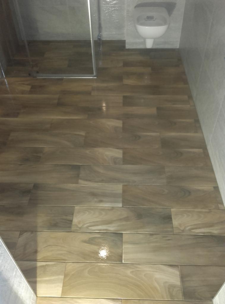 Ukończona podłoga w łazience wyłożona brązowymi, podłużnymi płytkami imitującymi drewno, z widocznym fragmentem kabiny prysznicowej i toalety.