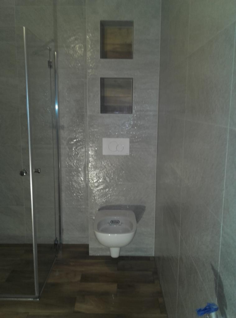 Wykończona łazienka w Skawinie: szklana kabina prysznicowa, podwieszany sedes i dekoracyjne wnęki w szarej, płytkowej ścianie nadają wnętrzu nowoczesny charakter.