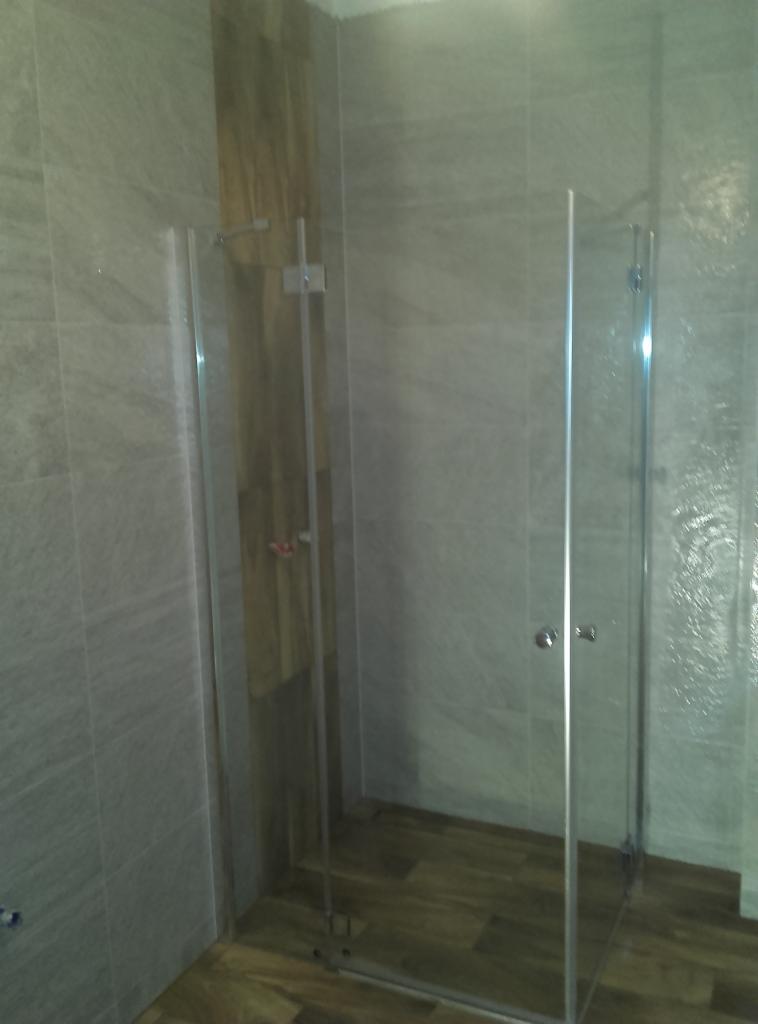 Nowoczesna kabina prysznicowa z przezroczystego szkła, wykończona w szaro-brązowej tonacji, z widocznym wykończeniem podłogi imitującym drewno.