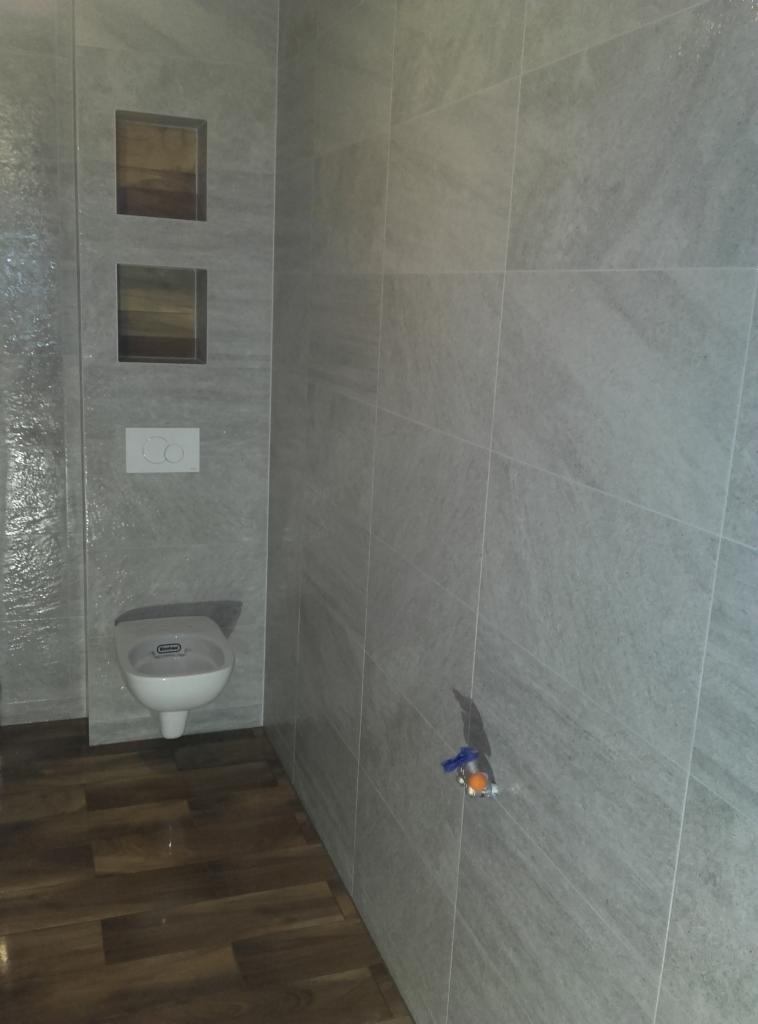 Wykończona łazienka z szarymi płytkami imitującymi kamień, drewnopodobną podłogą i podtynkową toaletą. Widoczne wnęki w ścianie i zawór wody.