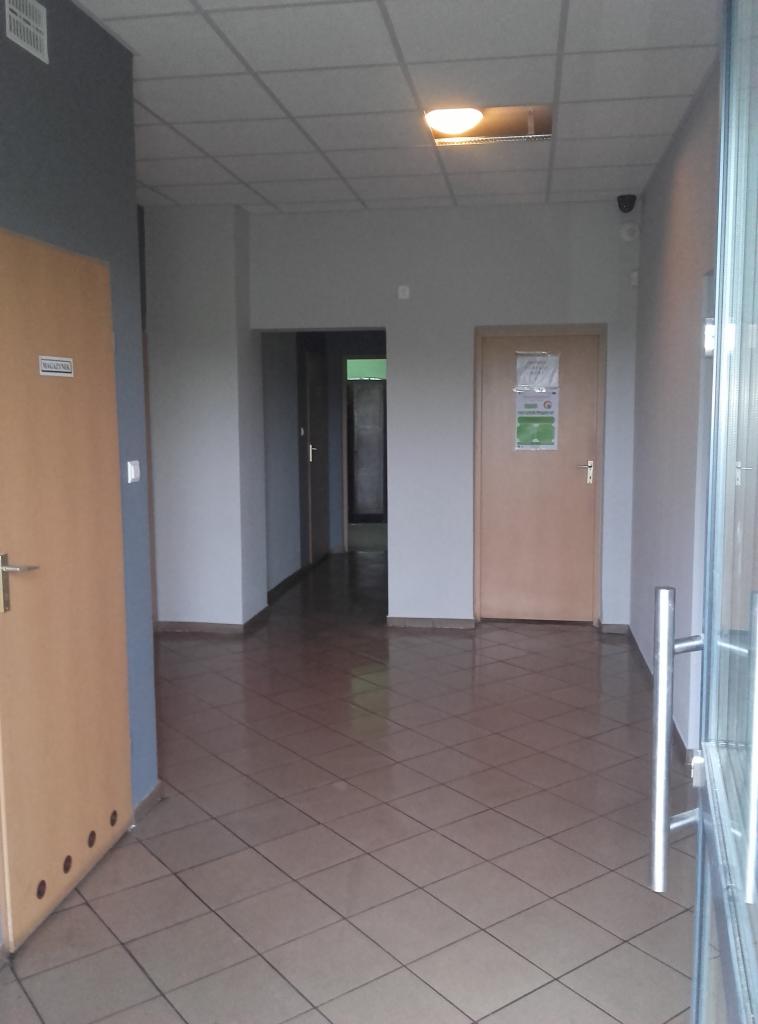 Korytarz biurowy z brązową podłogą w kratę, jasnymi ścianami i drewnianymi drzwiami. Widoczna lampa sufitowa i przeszklone wejście.