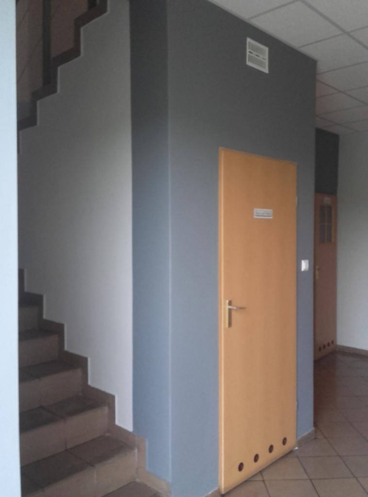 Korytarz po malowaniu: szare ściany, drewniane drzwi z metalową klamką, schody. Widoczne detale wykończenia i odświeżone kolory.