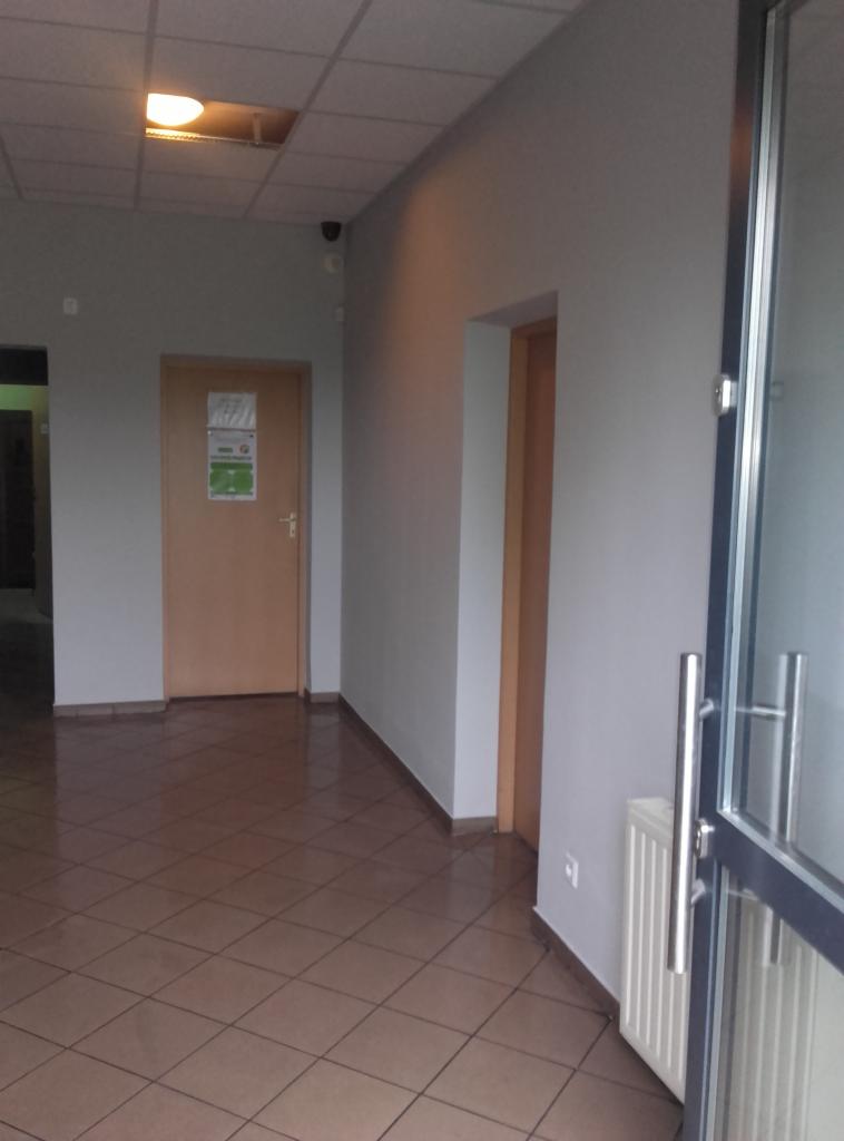 Korytarz biurowy z brązowymi płytkami podłogowymi w kratkę, szare ściany, drzwi z tabliczką informacyjną i częściowo widoczne szklane drzwi wejściowe.