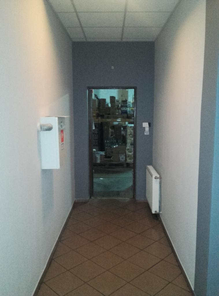 Wnętrze korytarza z brązową podłogą w romby, białą gaśnicą na ścianie i grzejnikiem. W tle widoczne otwarte drzwi do magazynu.
