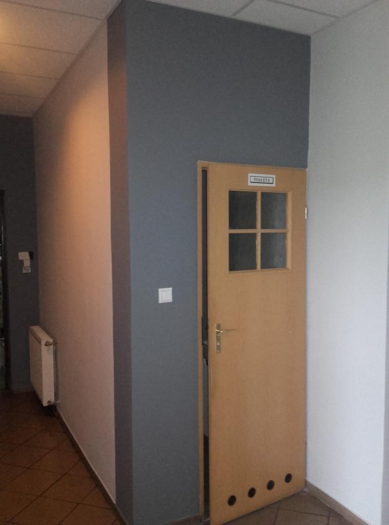 Ściana pomalowana na dwa kolory, szary i jasny beż, z drewnianymi drzwiami do toalety po prawej stronie. Widoczny fragment korytarza z grzejnikiem.