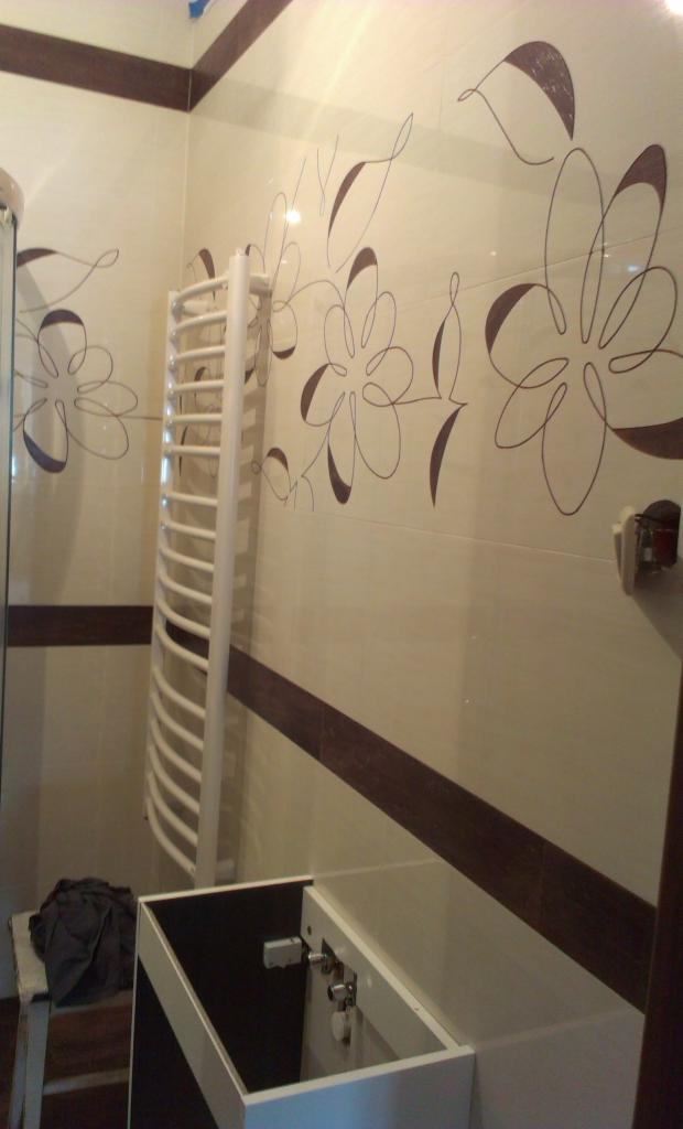 Łazienka w trakcie wykańczania: beżowe płytki z brązowym motywem kwiatowym, biały grzejnik drabinkowy i szafka pod umywalkę bez blatu, widoczne przyłącza.