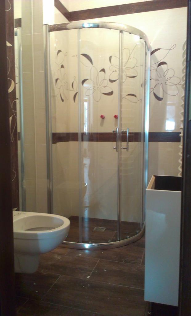 Wykończona łazienka z kabiną prysznicową z motywem kwiatowym, brązowymi płytkami podłogowymi i białym bidetem. Widoczna część sufitu z drewnianymi belkami.