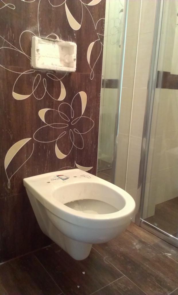 Biały bidet wiszący na ścianie z brązowymi płytkami w kwiaty, obok kabina prysznicowa. Widoczna puszka elektryczna. Wykończenie łazienki w trakcie.