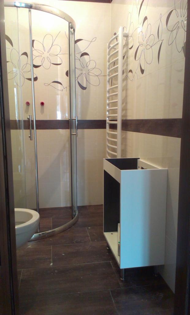 Wykończona łazienka z kabiną prysznicową, grzejnikiem drabinkowym i szafką pod umywalkę. Na ścianach kremowe płytki z brązowymi kwiatowymi motywami i pasem.