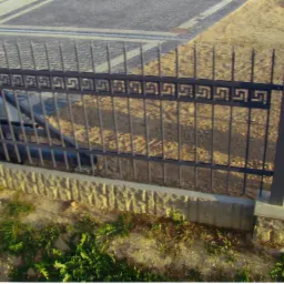 Fragment metalowego ogrodzenia z motywem meandrycznym, osadzonego na betonowej podmurówce wykończonej kamieniem dekoracyjnym, z widocznymi elementami automatyki bramy w tle.