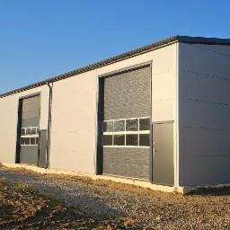 Nowo wybudowana hala z szarych płyt warstwowych z trzema bramami segmentowymi z oknami, widok z zewnątrz na teren budowy w słoneczny dzień.