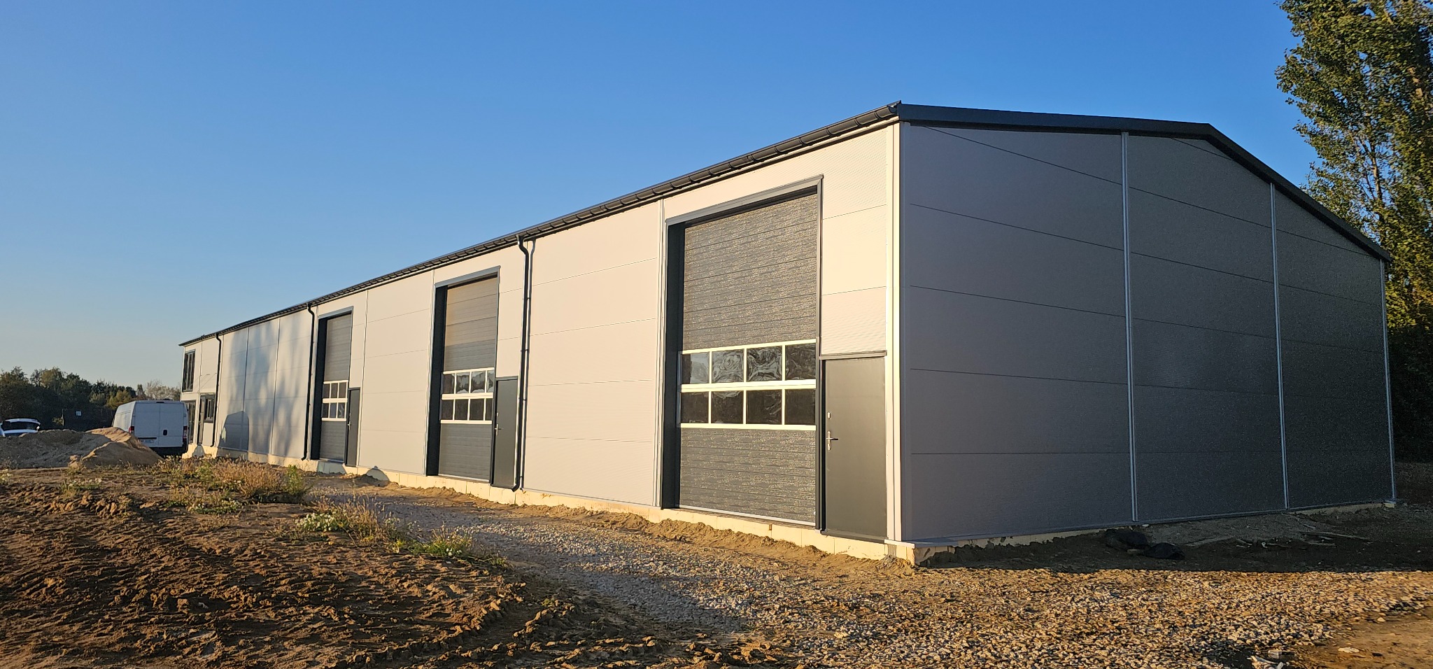 Nowo wybudowana hala z szarych płyt warstwowych z trzema bramami segmentowymi z oknami, widok z zewnątrz na teren budowy w słoneczny dzień.