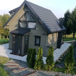 Fhu Łukasz Słowik - Nowoczesny, szary dom szkieletowy z czarnym dachem i numerem 694, otoczony zielenią, z widocznymi schodkami z desek i kamieni prowadzącymi do wejścia.