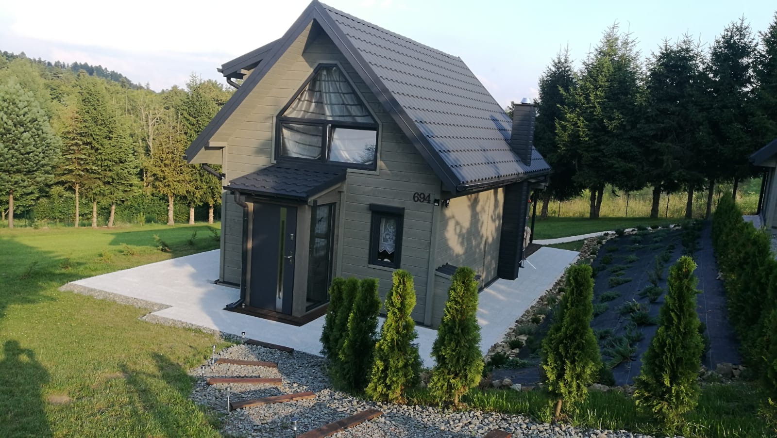 Nowoczesny, szary dom szkieletowy z czarnym dachem i numerem 694, otoczony zielenią, z widocznymi schodkami z desek i kamieni prowadzącymi do wejścia.