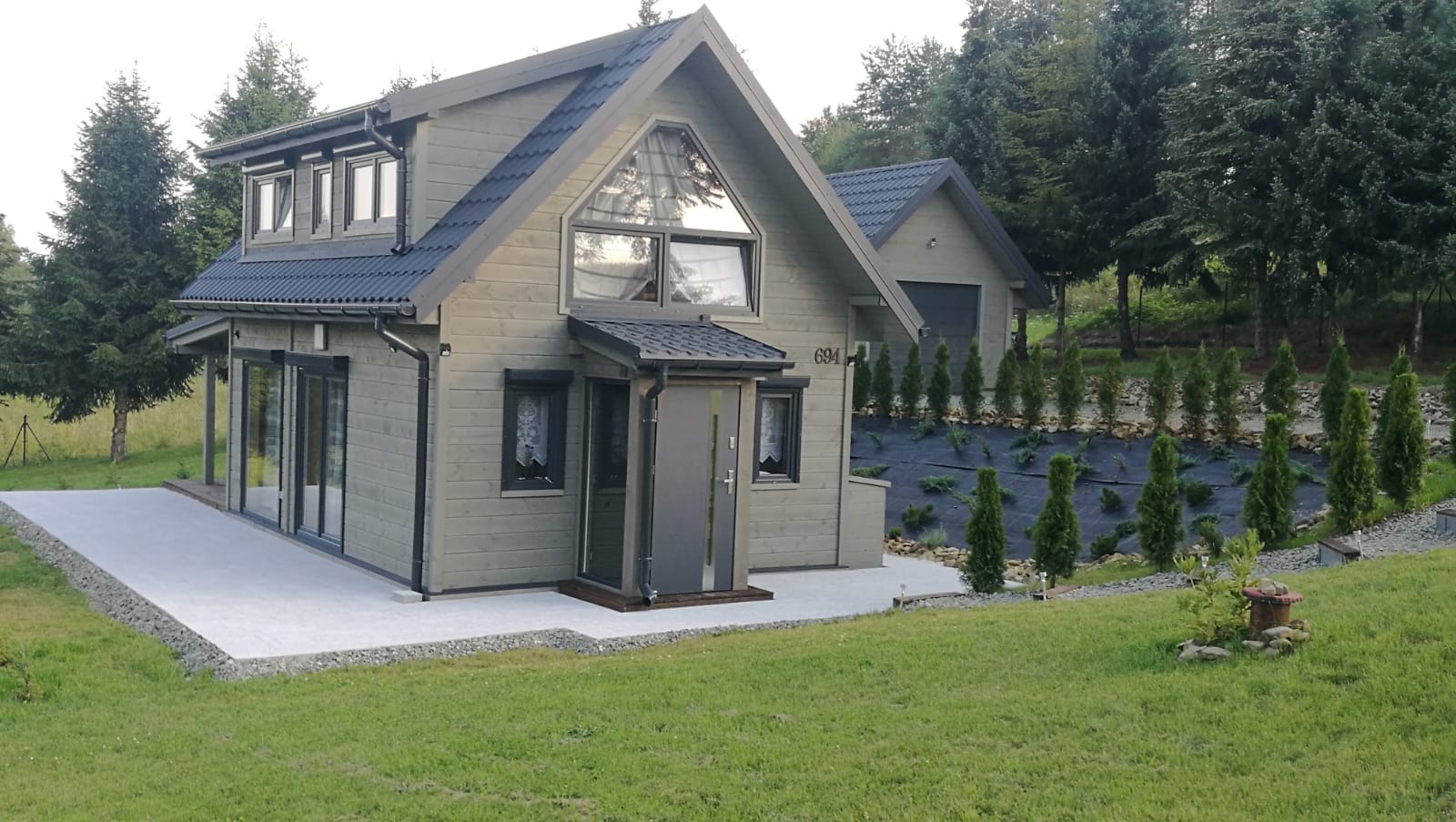 Nowoczesny dom z drewna w odcieniach szarości z dużymi oknami, wykończony z dbałością o detale, otoczony zielonym trawnikiem i ozdobnymi tujami.