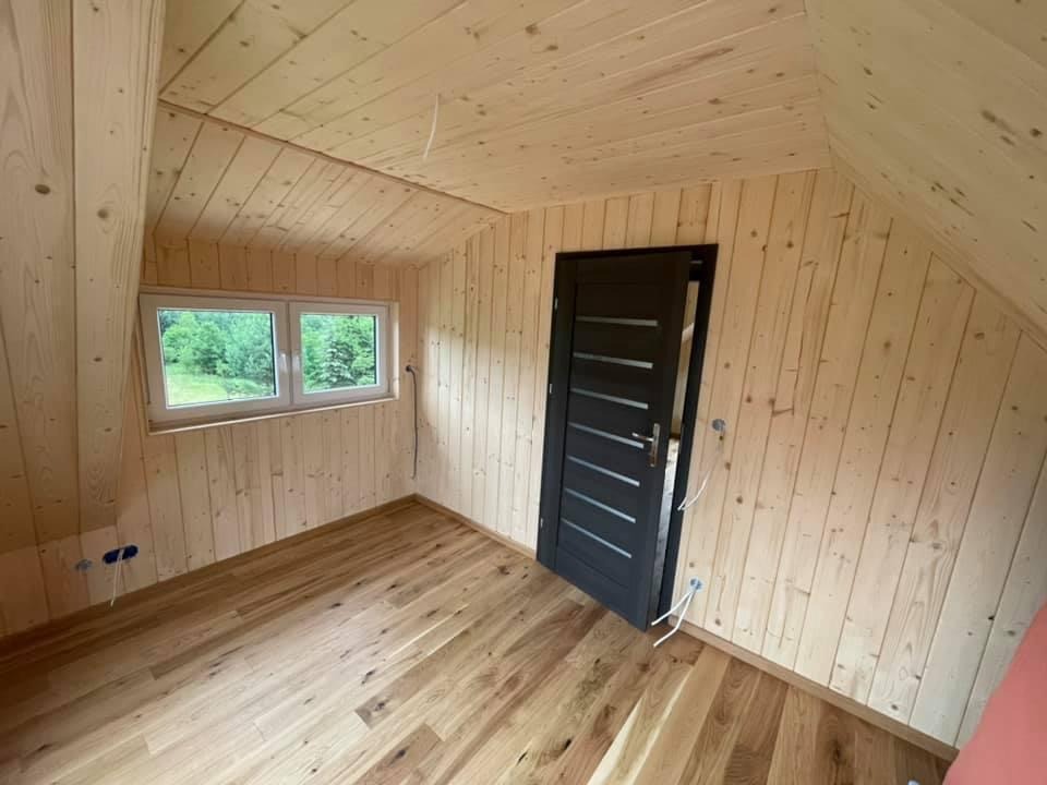 Wnętrze pokoju na poddaszu wykończonego boazerią i drewnianą podłogą, widoczne okno z widokiem na zieleń i czarne drzwi z poziomymi przeszkleniami.