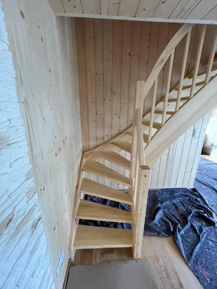 Wnętrze domu w trakcie wykańczania: drewniane schody z jasnego drewna, surowe deski na ścianach i suficie, zabezpieczona podłoga, widoczne ślady prac budowlanych.