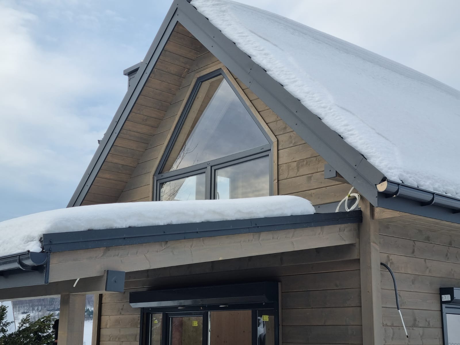 Fragment elewacji domu z bali w Starym Sączu, z trójkątnym oknem na poddaszu i śnieżną czapą na dachu.