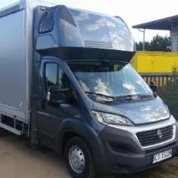 Szary bus Fiat Ducato z plandeką na podwórku, tablica rejestracyjna CT 1522M. Widoczne budynki w tle i ogrodzenie z siatki.