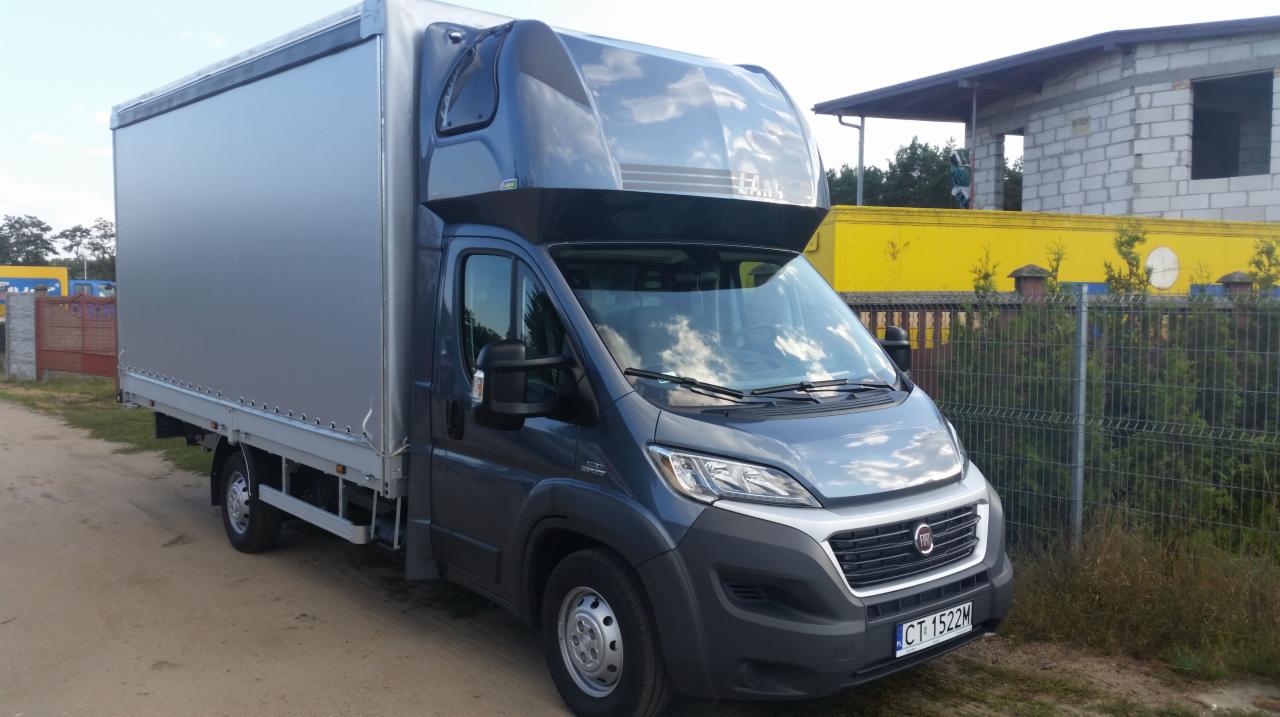 Szary bus Fiat Ducato z plandeką na podwórku, tablica rejestracyjna CT 1522M. Widoczne budynki w tle i ogrodzenie z siatki.