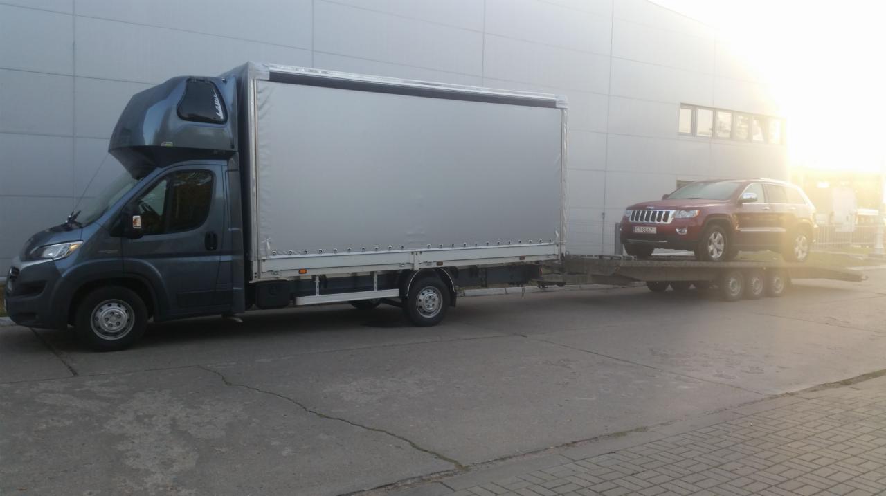 Szary bus z plandeką i kabiną sypialną transportuje na lawecie czerwonego Jeepa Cherokee na tle szarego budynku. Słońce w prawym górnym rogu.