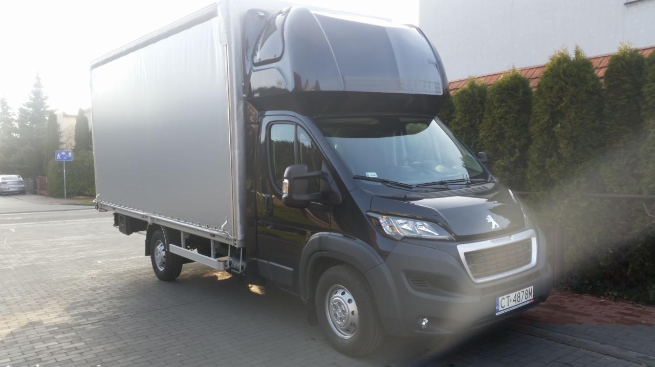 Czarny bus Peugeot z plandeką, zaparkowany na kostce brukowej. Widoczny numer rejestracyjny CT 4878M. W tle żywopłot z tui i budynek.