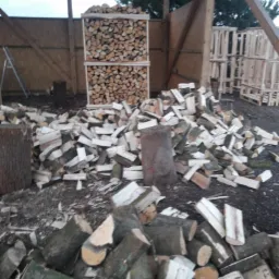 Stos drewna kominkowego w drewnianej ramie i luźno ułożone kawałki drewna na ziemi pod zadaszeniem.