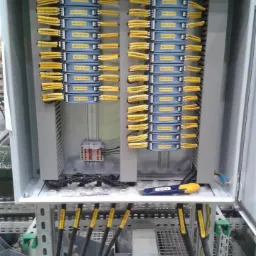 Otwarta szafa sterownicza z modułami i okablowaniem oznaczonym żółtymi etykietami. Widoczne narzędzie na dolnej półce. Szara obudowa, instalacja na metalowej konstrukcji.