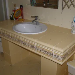 Beżowa umywalka wpuszczana w blat z płytek ceramicznych z dekoracyjnym paskiem, bateria chromowana, na blacie lusterko i kosmetyki, w tle płytki ścienne w delikatny wzór.