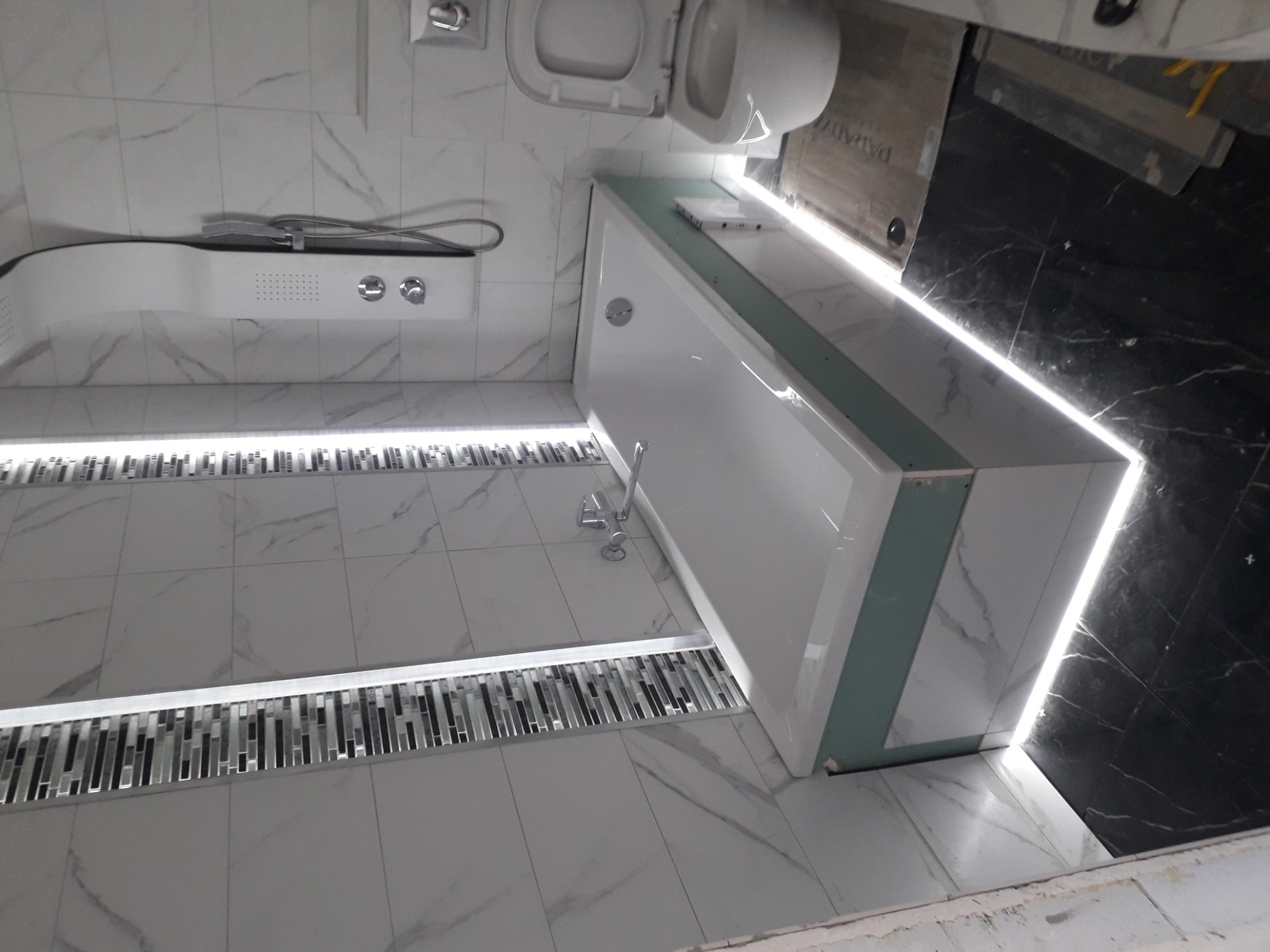 Nowoczesna łazienka z białymi płytkami imitującymi marmur, podświetlanymi listwami przypodłogowymi z mozaiki, białą wanną z zielonym obramowaniem i czarną ścianą w tle.